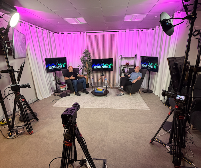 The Studio at River House: two hosts recording in armchairs against soft pink-lit curtains, with three cameras and three screens facing the set