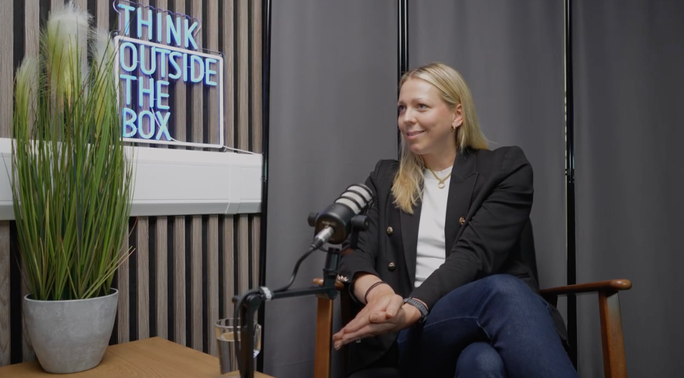 Ask The Arb podcast: a guest smiling in an armchair beside a microphone, with a Think Outside The Box neon sign behind